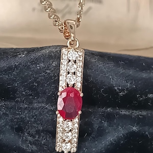 May include: A rose gold pendant necklace. The pendant has a central oval ruby, surrounded by small, clear gemstones. The rectangular pendant hangs from a rose gold chain. The necklace is displayed on a dark blue surface.