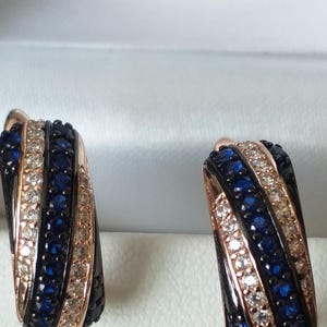 Könnte beinhalten: Ein Paar Creolen mit schwarzer und roségoldener Oberfläche. Die Ohrringe sind mit blauen und weißen Steinen verziert.