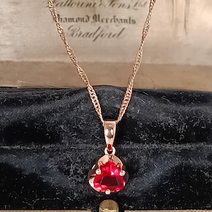 Könnte beinhalten: Eine roségoldene Halskette mit einem roten Edelsteinanhänger. Der Anhänger hat eine dreieckige Form mit facettierter Oberfläche. Die Kette ist im zarten Seil-Stil gehalten. Der Hintergrund enthält den Text "Tatterini & Sons Ltd Diamond Merchants Bradford".