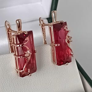 Puede incluir: Un par de pendientes de oro rosa con piedras rectangulares de color rojo intenso facetadas. Cada pendiente está decorado con un delicado diseño de enredadera metálica que incluye una pequeña piedra clara brillante en el lateral.