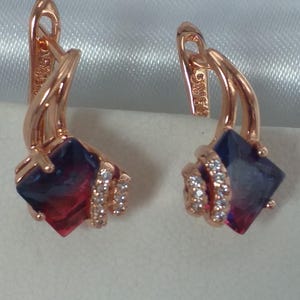 Op de afbeelding: Rosegouden oorbellen met een vierkante, tweekleurige edelsteen, die overloopt van rood naar blauw. Kleine, heldere stenen zijn rond de basis van de vierkante edelsteen gezet. De oorbellen worden gepresenteerd op een witte ondergrond.
