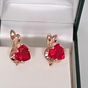 Op de afbeelding: Een paar roségouden oorbellen met hartvormige, gefacetteerde rode edelstenen. De oorbellen worden gepresenteerd in een wit gevoerde, groen omrande sieradendoos.