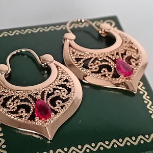 Op de afbeelding: Een paar roségouden oorbellen met ingewikkelde filigraan details. Elke oorbel heeft een druppelvormige, helderrode edelsteen. De oorbellen worden gepresenteerd op een donkergroene doos met een decoratieve rand.