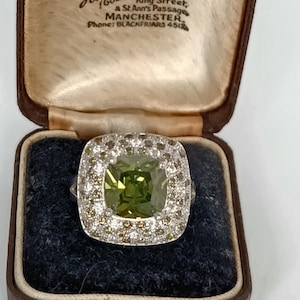 Può includere: Un anello d'argento con una gemma quadrata verde oliva, circondata da un alone di diamanti bianchi e marroni. L'anello è presentato in un portagioie vintage marrone. Il portagioie ha un testo che recita "Hancock & Son Ltd, Diamond Merchants, Manchester".