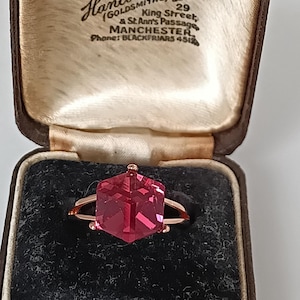 May include: A rose gold ring with a large, faceted, hexagonal, red gemstone. The ring is displayed in a brown jewelry box with the text "Hancock & Son" and "Diamond Merchants" visible on the inside of the lid.