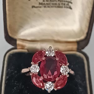 Peut inclure: Une bague ornée d'une grande pierre rouge centrale de taille ovale, entourée de quatre pierres rouges taille marquise et de quatre pierres blanches transparentes taille ronde, présentée dans un écrin marron avec du texte imprimé.