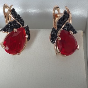 May include: A pair of rose gold earrings featuring a teardrop-shaped red gemstone and black detailing. The earrings have a lever back and are presented on a white surface.