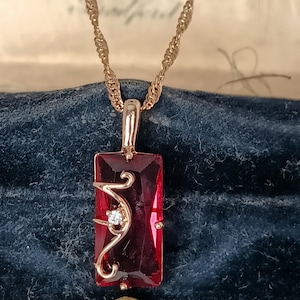 Op de afbeelding: Een rechthoekige rode edelsteen hanger hangend aan een goudkleurige gedraaide ketting. De hanger is voorzien van een gefacetteerde dieprode steen met een goudkleurige krulversiering en een kleine heldere steen in het metaal.