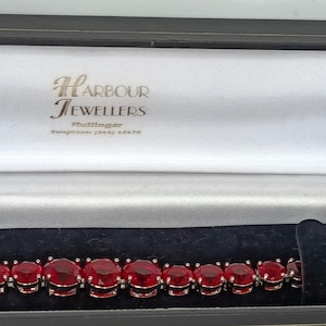 May include: A tennis bracelet featuring a series of round, faceted, deep red stones set in silver-toned metal. The bracelet is displayed inside a black and white jewelry box with the text Harbour Jewellers Mullingar printed on the interior lid.