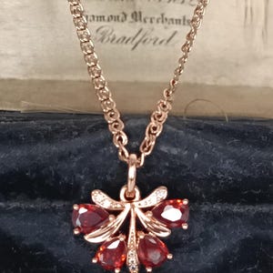 May include: A rose gold necklace with a pendant featuring a cluster of red gemstones and small white diamonds. The pendant is shaped like a stylized flower or bow. The chain is delicate and the necklace is displayed in a dark blue velvet box.