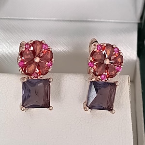 Peut inclure: Une paire de boucles d'oreilles en or rose. Chaque boucle d'oreille présente un motif de fleur rouge foncé avec de petites pierres roses et une pierre carrée, violet foncé. Les boucles d'oreilles sont présentées sur une surface blanche.