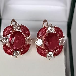 Puede incluir: Un par de pendientes ovalados con una piedra roja central rodeada por un halo de piedras rojas talla marquesa y pequeñas piedras claras redondas, todo montado en metal rosado con cierres de palanca para orejas perforadas.