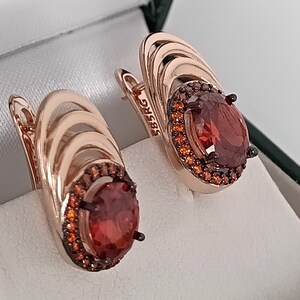 Può includere: Un paio di orecchini in oro rosa con pietre preziose rosse ovali. Gli orecchini hanno un alone di pietre più piccole rosso-arancio. Il metallo è timbrato con "585RG". Presentati in una scatola bianca.