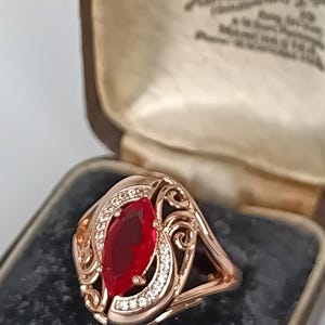 Può includere: Un anello in oro rosa con una gemma rossa centrale taglio marquise circondata da una fila curva di piccole pietre chiare. La fascia in metallo presenta intricati dettagli a volute e filigrana, presentato all'interno di un portagioie.