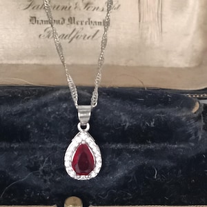 May include: Silver pendant necklace with a teardrop-shaped, deep red gemstone, encircled by small, clear stones. The pendant hangs from a delicate chain. The necklace is displayed in a vintage jewellery box with the text "Tattersall & Sons Diamond Merchants Bradford".
