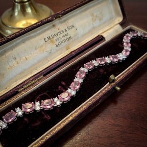 Pode incluir: Uma pulseira de tom prateado com uma série de pedras preciosas cor-de-rosa de corte oval alternadas com pequenas pedras claras de corte redondo. A pulseira é exibida dentro de um estojo alongado, articulado e forrado a veludo roxo com o texto E.M. DAVIES & SON. EST. 1892. LONDON.