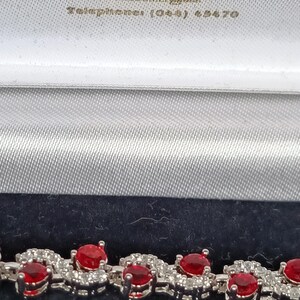 May include: A silver-toned bracelet featuring a repeating S-shaped link design, each set with a round, vibrant red gemstone and accented with small, clear pavé-set stones. The bracelet is presented in a white and dark blue jewellery box.