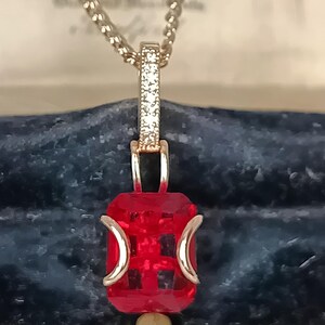 Könnte beinhalten: Eine silberne Halskette mit einem rechteckigen, facettierten, roten Edelsteinanhänger. Der Anhänger ist mit goldfarbenem Metall akzentuiert und ein kleiner Teil der Kette ist mit kleinen, klaren Steinen besetzt. Die Halskette wird vor einem dunkelblauen Hintergrund präsentiert.