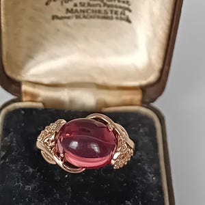 Puede incluir: Un anillo de oro con una gran piedra preciosa rosa de corte cabujón ovalado, engastada en una banda de metal decorativa. El anillo se muestra dentro de un joyero marrón con el texto "Hancock & Son, Goldsmiths, 29 King Street, St Ann's Passage, MANCHESTER".