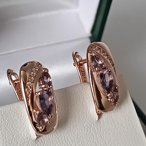 Peut inclure: Une paire de boucles d'oreilles en or rose avec un design incurvé. Chaque boucle d'oreille comporte trois pierres précieuses violettes et de petites pierres claires. Les boucles d'oreilles sont présentées sur une surface blanche à l'intérieur d'une boîte blanche.