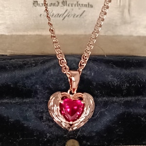 Op de afbeelding: Rosegouden hartvormige hangerketting met een robijnrode edelsteen in het midden en vleugelachtige accenten. De ketting heeft een delicate rolo-stijl. De ketting wordt gepresenteerd in een vintage sieradendoos. De doos heeft de tekst "Fadorini & Sons Ltd Diamond Merchants Bradford."