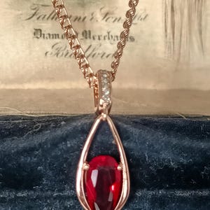 May include: A rose gold-toned necklace featuring a pear-shaped red gemstone pendant suspended within an open teardrop frame. The pendant bail is accented with small clear stones, and the chain is a twisted rope style. The metal has a warm pink hue.