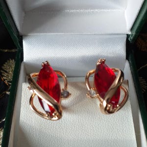 Op de afbeelding: Een paar gouden oorbellen met rode, markiesvormige edelstenen en kleine, heldere stenen. De oorbellen bevinden zich in een groene juwelendoos.