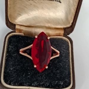 May include: A rose gold ring featuring a large, faceted, marquise-cut red gemstone. The ring is presented in a vintage brown jewellery box. The box has the text "Hancock & Son Ltd" and "Manchester".