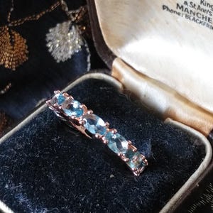 May include: A ring with a row of oval-shaped blue gemstones set in a rose gold band. The ring is in a brown velvet box with a white satin lining. The box has a label that reads "Diamond Merchants Hancock & (Goldsmith King & St. Ann's Manche Phone: Blackfriars".