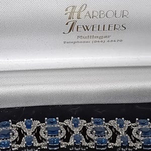 Puede incluir: Una pulsera de tono plateado con piedras azules de corte ovalado rodeadas de pequeñas piedras transparentes, presentada en un joyero blanco con el texto Harbour Jewellers y un número de teléfono impreso en el forro interior.