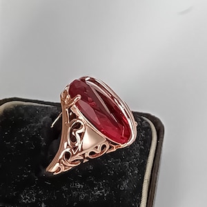 Puede incluir: Anillo de oro rosa con una gran gema roja de corte ovalado. El anillo tiene un diseño de filigrana ornamentado en la banda y alrededor del engaste de la gema. El anillo se exhibe sobre una superficie de terciopelo negro.
