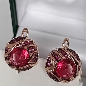 Pode incluir: Um par de brincos redondos com uma grande pedra vermelha facetada no centro, rodeada por uma armação de metal em tom rosado com detalhes em espiral e pequenos cristais rosa-escuros incrustados. Fecho de alavanca seguro.