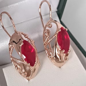 May include: A pair of rose gold earrings with a red, teardrop-shaped gemstone. The earrings feature an open filigree design around the gemstone and a hook closure. The earrings are displayed in a white jewelry box.