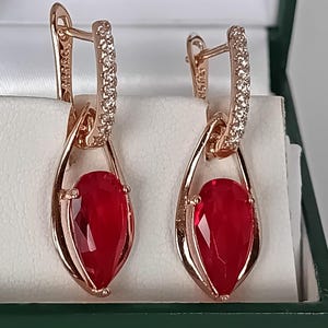 Puede incluir: Un par de pendientes colgantes de oro rosa con una piedra roja en forma de pera suspendida dentro de un marco en forma de lágrima. El aro superior de cada pendiente está incrustado con pequeñas piedras claras y brillantes que contrastan.