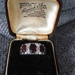 May include: A silver ring with three oval, deep red gemstones, flanked by smaller clear stones, set in a vintage brown jewelry box. The box is lined with cream satin and black velvet. The box has the text "DIAMOND MERCHANTS" and "Hancock & Son".