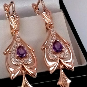May include: A pair of rose gold dangle earrings with a purple gemstone and clear cubic zirconia accents. The earrings are in a box.