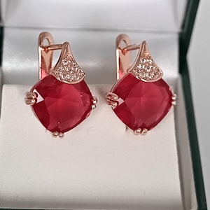 Puede incluir: Un par de pendientes de oro rosa con grandes piedras preciosas rojo rubí cuadradas. La parte superior de cada pendiente presenta un diseño triangular incrustado con pequeños cristales transparentes. Los pendientes se muestran en una caja de joyería verde y blanca.
