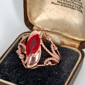 Puede incluir: Un anillo de oro rosa con una gran gema roja de talla ovalada. El anillo presenta intrincados detalles de cuerda y cuentas alrededor de la piedra central. El anillo se muestra en un joyero vintage.