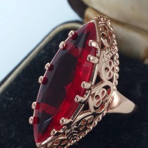 May include: A rose gold ring with a large, faceted, deep red gemstone. The ring features an ornate filigree design around the stone and on the band. The gemstone is approximately 1.5 inches long.