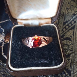 May include: A gold ring with a large, red gemstone and smaller, clear gemstones set in the band. The ring is in a brown velvet box with a white label that reads "Hancock & Son (Goldsmiths) Ltd. 29 King Street & St. Ann's Passage Manchester Phone: BLACKFRIARS 4512".