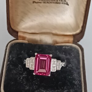 Op de afbeelding: Een zilverkleurige ring met een grote, rechthoekige, smaragdgeslepen roze edelsteen in het midden, geflankeerd door twee kleinere rechthoekige secties bezet met kleine heldere steentjes. De ring wordt gepresenteerd in een bruin juwelendoosje met tekst.