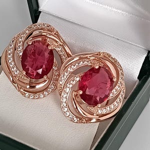 Op de afbeelding: Een paar roségouden oorbellen met grote ovale, robijnrode edelstenen. De oorbellen hebben een wervelend ontwerp met kleine, heldere kristallen. Ze worden gepresenteerd in een zwart-witte sieradendoos.