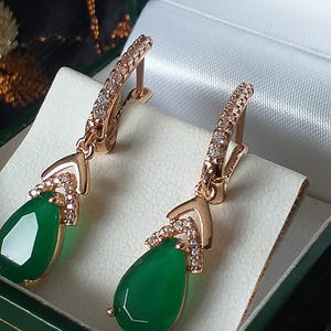 Può includere: Un paio di orecchini in oro rosa con gocce di pietre preziose verdi e accenti di zircone cubico trasparenti. Gli orecchini sono esposti in una scatola di velluto verde.
