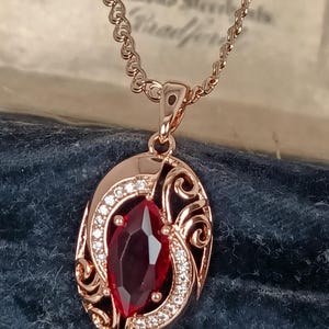 May include: A rose gold-toned pendant necklace featuring an oval frame with intricate scrollwork and small clear stones. A large, marquise-cut deep red gemstone is set in the center. The pendant hangs from a matching rose gold-toned chain.