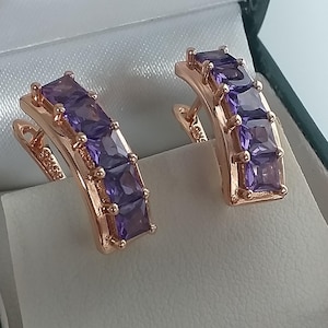 May include: Rose gold earrings, each featuring three square, purple gemstones. The earrings are presented in a white-lined jewellery box with a dark green border.