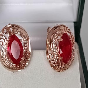 Pode incluir: Um par de brincos em ouro rosa com uma pedra preciosa vermelha de corte marquise ao centro, rodeada por uma estrutura curva com volutas intrincadas e pequenas pedras transparentes cravadas em pavé. Acabamento metálico polido.