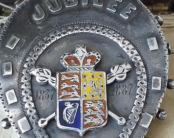 Może przedstawiać: Srebrna broszka w kształcie podkowy z herbem królewskim w środku. Broszka jest ozdobiona napisem "JUBILEE" oraz datami "1837" i "1887".