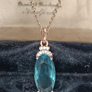 May include: A rose gold necklace featuring a large, oval, teal gemstone pendant. The pendant is adorned with small, clear crystals. The chain has a delicate, twisted design.