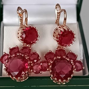 Puede incluir: Un par de pendientes colgantes de oro rosa con una piedra grande, redonda y facetada de color rojo intenso, rodeada de piedras rojas circulares más pequeñas en un diseño de racimo. Cada pendiente tiene una piedra roja redonda arriba y cierre.