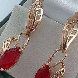 Könnte beinhalten: Ein Paar goldfarbene Ohrringe mit einem blattartigen Design. Jeder Ohrring hat einen tiefroten, marquiseförmigen Edelstein. Die Ohrringe werden in einer dunkelgrünen Schmuckschatulle mit weißem Innenraum präsentiert.